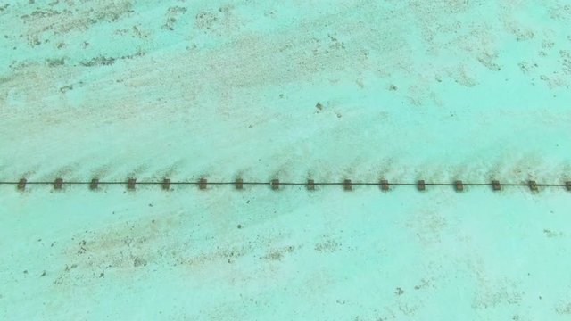DRONE, TOP DOWN: Flying above a sewage drainpipe running across a white sand shore and underneath the beautiful turquoise ocean water. Waste conduit runs across the shallows near an island in Maldives