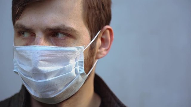 Sick infected bearded man with green eyes wear protective disposable medical face mask, stares longingly at the camera close-up. Portrait of ill beautiful guy on gray background. Isolation concept.