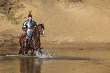 A young adult man in knightly armor rides a horse on a river along a sandy shore
