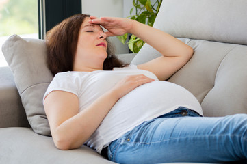 Pregnant woman with hand on forehead suffering headache lying on a couch in the living room at home