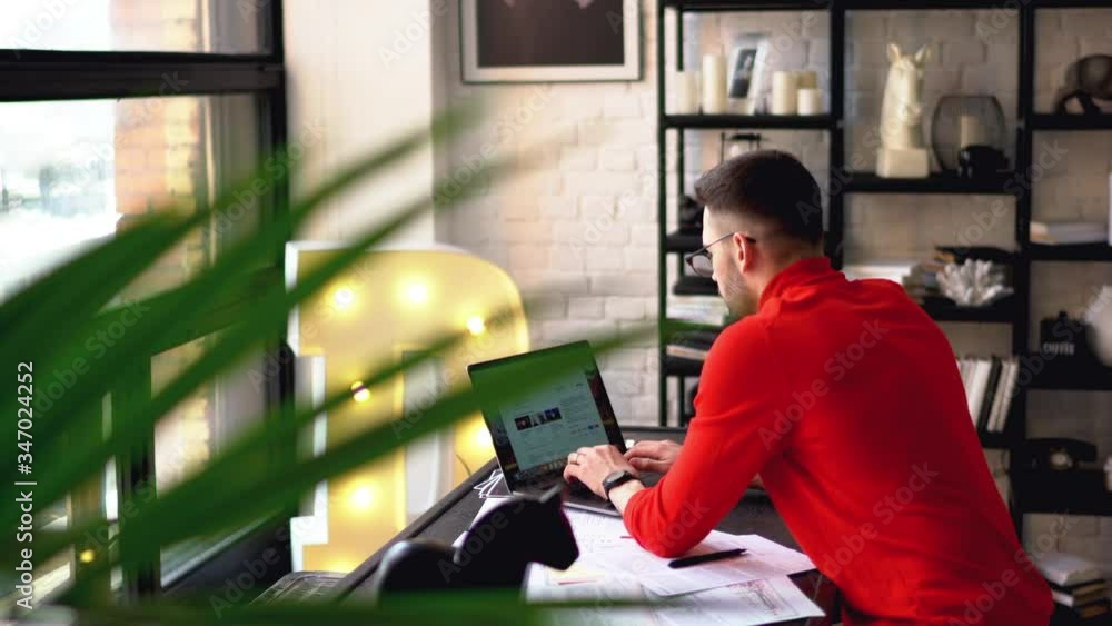 Poster young man in eyeglasses for vision correction working remotely at modern laptop to create design on 
