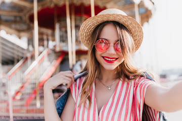 Refined young lady in elegant hat making selfie in front of carousel. Outdoor portrait of debonair caucasian girl spending weekend in amusement park. - obrazy, fototapety, plakaty