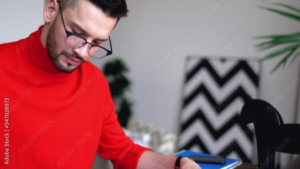 Poster pensive young man writing notes on paper sketches sitting at desktop with laptop in modern flat.bear