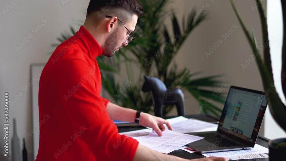 Poster smart young bearded man in casual wear checking paper documents and preparing financial report at mo