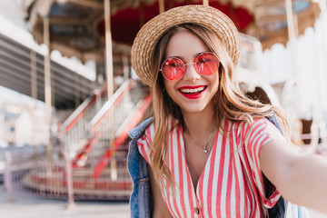 Pleased white girl in round glasses making selfie near carousel. Outdoor shot of cheerful blonde woman in straw hat taking picture of herself in amusement park. - obrazy, fototapety, plakaty