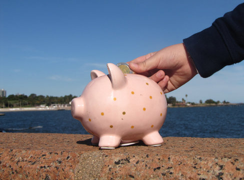 Money saving in piggy bank. A young boy puts a coin in a pink ceramic piglet piggyback on the waterfront with the city and the sea in the background lit by sunlight