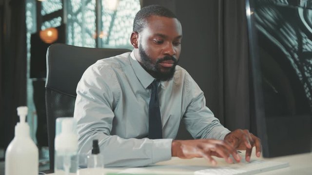 African American handsome man uses computer for work in modern office programmer serious looks at monitor screen workplace engineering problems slow motion