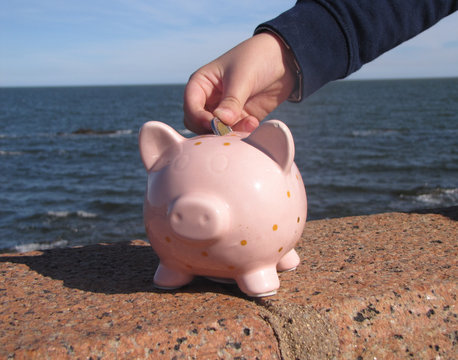 Money saving in piggy bank. A young boy puts a coin in a pink ceramic piglet piggyback on the waterfront with the city and the sea in the background lit by sunlight