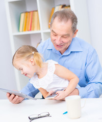 Senior and a little girl sitting on his lap are sitting in a room. Grandfather holding a tablet in his hand and looking at granddaughter. Little girl holding a mobile phone in her hands