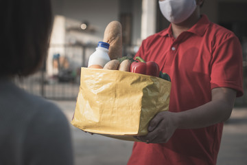 Deliver man wearing face mask in red uniform handling yellow bag of food, fruit, milk, vegetable give to female costumer Postman and express grocery delivery service during covid19.