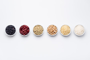 Rice and beans bowls top view. Uncooked staple food ingredients like chickpeas, red kidney beans, green lentils, black turtle beans, jasmine rice and basmati rice on white background from above.