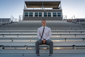 A teenage boy, a senior in high school  class of 2020 sits alone on the high school stadium bleachers.