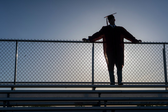 A graduating high school teenage boy wearing a cap and gown leans against a stadium fence and is silhouetted by the setting sun. 