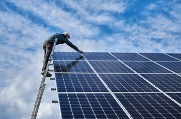 Male worker electrician repairing photovoltaic solar module, standing on ladder under beautiful...