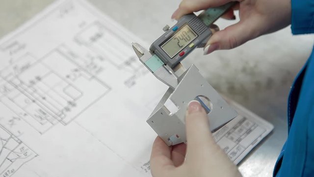 Engineer is measuring dimensions of detail with digital vernier caliper equipment and design drawing. Numbers on the electronic caliper display. High-precision equipment at factory.