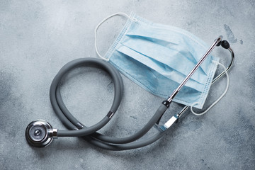 Stethoscope and protective medical face mask on a light-grey stone background, view from above, horizontal shot