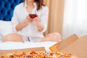 Pepperoni pizza, against the background of a girl in a white shirt sitting on bed with glass red...