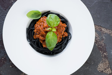 White plate with black spaghetti, bolognese sauce and fresh green basil, above view on a brown stone background