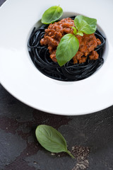 Close-up of squid-ink spaghetti with bolognese sauce and fresh green basil in a white plate, selective focus, vertical shot