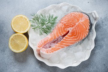 White shell-shaped plate with raw iced salmon steak, fresh dill and lemon over light-blue stone background, horizontal shot