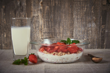 Muesli with yogurt and strawberries in a glass bowl, next to a glass of milk and a wooden spoon.
