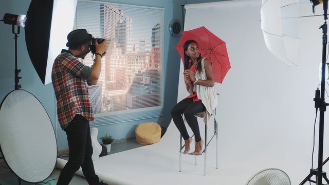 Stylish african model posing with red umbrella on bar high chair for fashion magazine photo shoot. Photographer taking photos on professional camera.