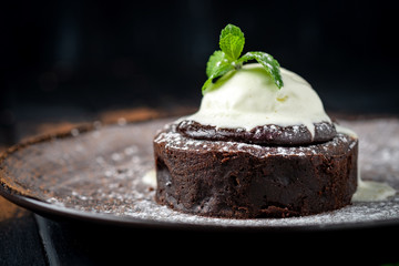 Chocolate brownie with a ball of melting vanilla ice cream, mint, powdered sugar and cocoa on a plate on a dark wooden background