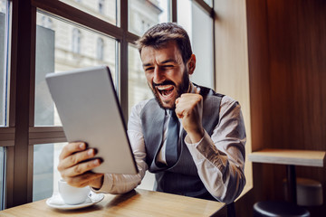 Fototapeta premium Young successful businessman sitting in coffee shop, looking at tablet and celebrating.