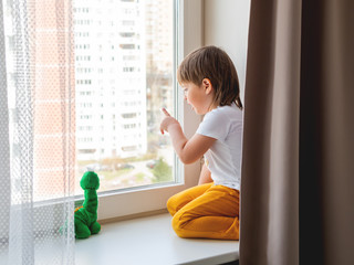 Toddler boy sits with green dragon or dinosaur. Kid with plush toy look through window outside. Child on home quarantine because of coronavirus COVID-19 waiting for somebody. © Konstantin Aksenov