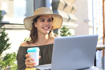 Happy nice woman working on laptop in street cafe, holding paper cup.