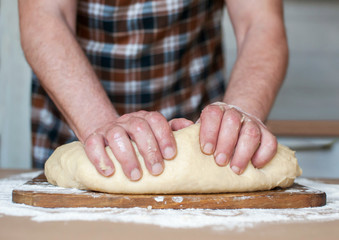 A man in an apron kneads the dough on a wooden board. Hands close up