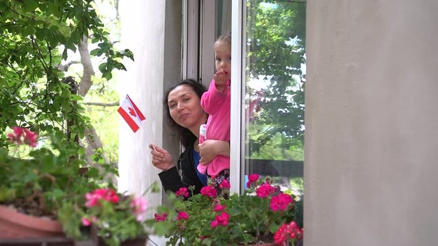 Canadian family mother and little girl child on balcony / window are smiling and waving the flag of Canada. Self-isolation in quarantine, lockdown, stay at home, social distancing, coronavirus