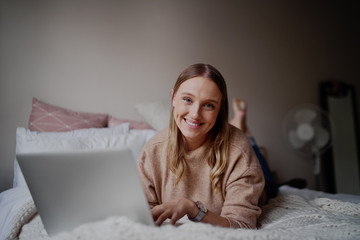 Cheerful female entrepreneur lying on her bed working online on her laptop in bedroom during self isolation - student learning online classes