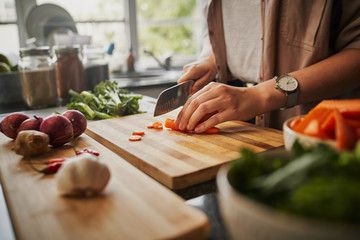 Closeup of young female hands chopping fresh orange carrot on board while in modern kitchen -...