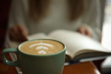 Woman with coffee reading book indoors, focus on cup