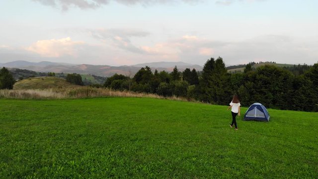Aerial drone view of young happy woman stands near tent and raises her hands up at background of scenic mountain sunset landscape.
