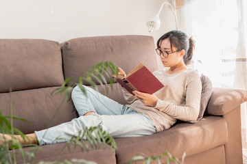 Asian woman relaxing at home reading an old book on the sofa. Girl enjoying quiet time indoors. lying on the couch.