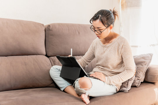 Young Asian modern woman working in graphic design and art from home. Girl sitting and drawing on an ipad tablet on the sofa during coronavirus pandemic quarantine.