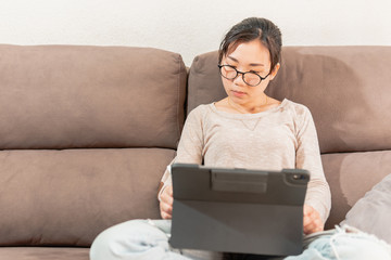 Young asian woman graphic designer drawing on an ipad tablet. Girl working from home sitting on the sofa couch. Work from home due to coronavirus pandemic.