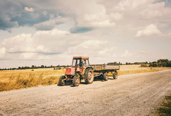 Fototapeta premium Old Red Tractor with a Trailer moving on the Countryside Sandy Road in hot summer day. Father and Son inside a tractor.