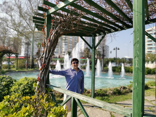 A middle-aged woman in sunglasses stands and smiles in a gazebo in the park against the background of a fountain.