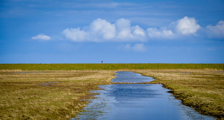 One man hiking with a backpack in the distance on the horizon at a wetland wildfowl reserve with a flood ditch lake receding almost to vanishing point, under a single line of forming cumulous clouds.