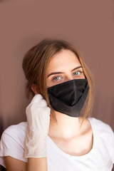 portrait of beautiful young woman wearing cotton black mask and white medical/surgical gloves on pastel background. She is adjusting mask. 