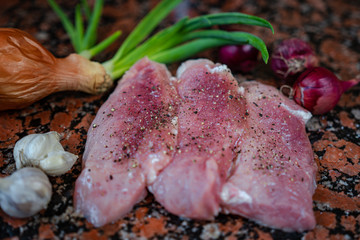 Raw pork with pieces of coarse salt and pepper on top of a marble table near sprouted onions. Fresh steaks on the table next to blue onions and garlic. Side view in natural light.