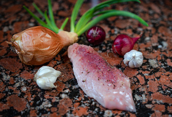 Raw pork with pieces of coarse salt and pepper on top of a marble table near sprouted onions. Fresh steaks on the table next to blue onions and garlic. Side view in natural light.