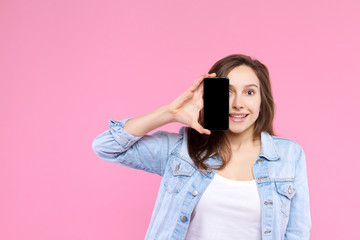 Pretty smiling girl is holding in hand smartphone with empty blank screen display. Young woman dressed in denim jacket and white t-shirt on pink background. Modern technology and devices concept.