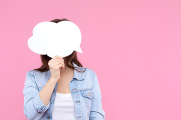 Girl in white t-shirt, denim jacket is holding in hand in front of head paper decorative thinking, speaking cloud. Young woman is standing on pink background. Emotional portrait concept.