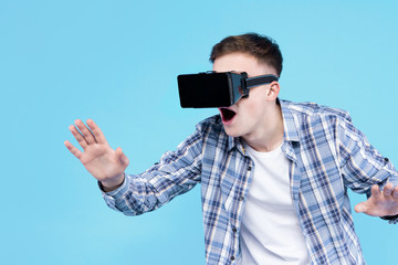 Shocked, surprised guy student in VR glasses on blue background. Young man dressed in checkered shirt, white t-shirt is enjoying virtual reality world. Modern technology and devices concept.