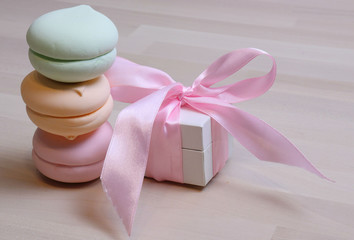 three multi-colored marshmallows stand in the form of a tower, on the wooden surface of the table. Nearby is a small present, a white box tied with a pink ribbon. Place for inscriptions, text. Holiday