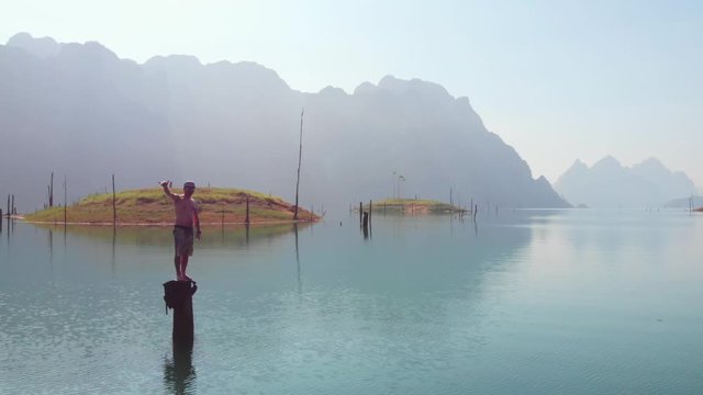 Man Standing On A Timber With Beautiful Water Reflections At The Middle Of Peaceful Clear Lake Under Brilliant Blue Sky In Thailand - Drone (Mavic)